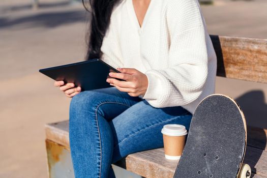 unrecognizable young woman sitting on a bench using her tablet next to a coffee and a skateboard
