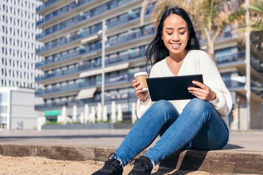 young asian woman smiling sitting on the city watching her tablet with a take-away coffee in her hand