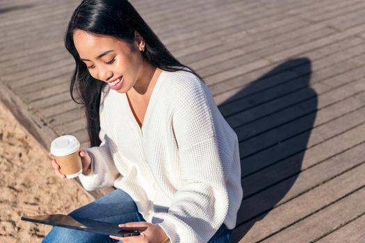 young asian woman smiling sitting outdoors looking at her tablet with a take-away coffee in her hand