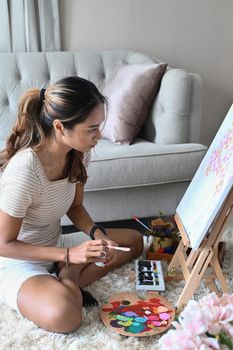 Side view millennial woman in casual clothes sitting on floor in living room and painting picture on canvas.