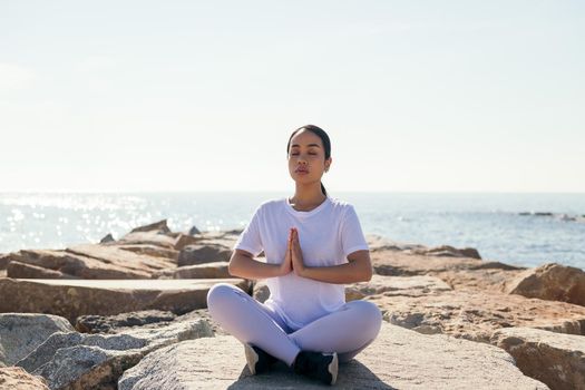 young asian woman relaxing doing a meditation with eyes closed and hands together as if praying sitting by the sea, concept of spirituality and relax