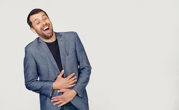 Young businessman man with a beard in a jacket smiling and laughing out loud because a funny crazy joke with hands on the body. Portrait of a man on a gray background.