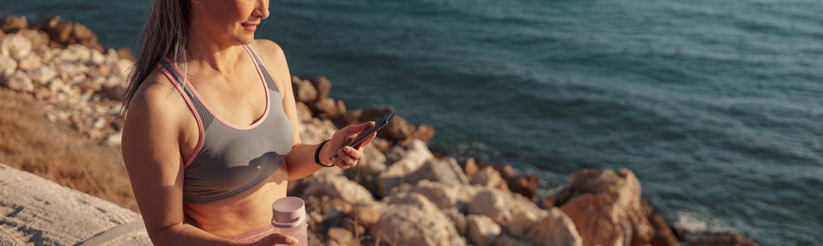 Slender adult woman in sports clothes sitting by the seashore, holding buttle of water and mobile phone