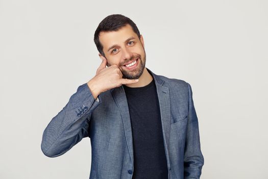 Young businessman man with a beard in a jacket, smiling, making a telephone gesture with his hand and fingers, like talking on the phone. Transfer of concepts. Portrait of a man on a gray background.