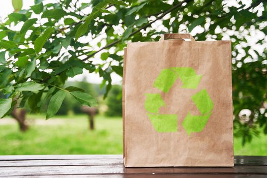 Blank paper bag stands on a wooden table against the background of green leaves. Shopping concept. High-quality photo.