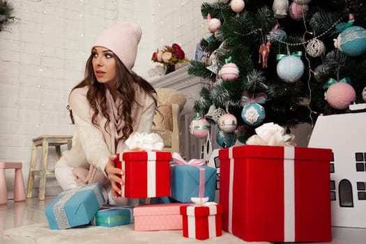 Happy young woman putting gift boxes under the Christmas tree. Gifts for the family. The girl in the sweater is preparing a surprise. Christmas New Year holiday concept