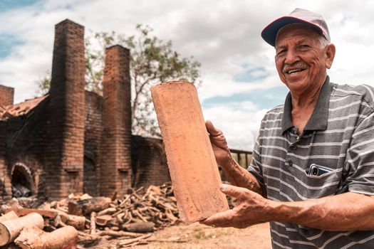 Elderly man in a hat smiling and looking at the camera holding a clay tile in his hands in front of a brick kiln in La Paz Centro Nicaragua
