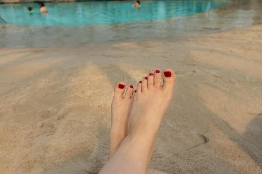 beautiful female feet with red pedicure on the background of blue water and sand on the beach. the concept of relaxation by the sea on a bright, hot and sunny day. soft focus