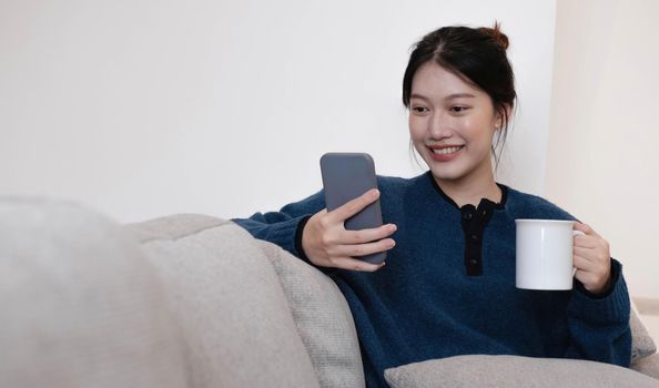 Portrait of a smiling asian women sitting at sofa and drink coffee looking at smartphone.