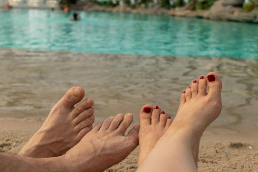 male and female feet lie on the sand against the background of blue water. two pairs of legs. concept of relaxation by the sea on a bright, hot and sunny day on the beach. soft focus