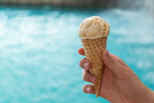 young hand holds an ice cream cone on the background of the blue water of the sea. the concept of relaxation by the sea on a bright, hot and sunny day. soft focus