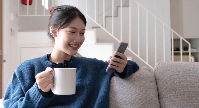 Portrait of a smiling asian women sitting at sofa and drink coffee looking at smartphone.