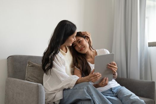 Happy young lesbian couple hugging having fun using digital tablet relaxing on couch at home. Two smiling asian women friends holding computer looking at screen enjoying surfing online watching videos