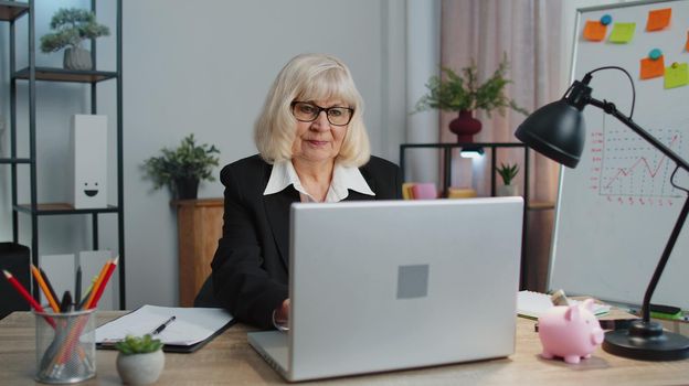 Smiling professional mature business woman sits at workplace desk, opening laptop computer, start working. Happy senior older employee 60s businesswoman executive working typing on pc at home office