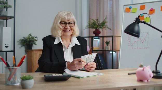 Senior business woman accountant or banker using calculator making calculations at workplace desk. Elderly grandmother counting money cash dollar banknotes working, writing on notebook at home office
