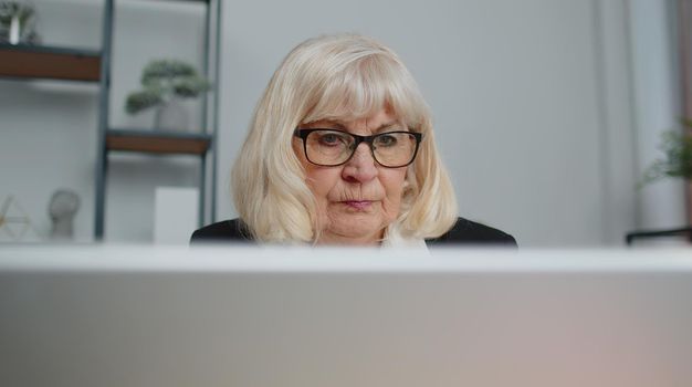 Close-up face professional senior business woman employee wears glasses using laptop computer sits at workplace. Elderly grandmother 70s freelancer executive working typing on keyboard at home office