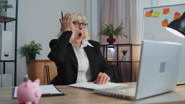 Oh my God Wow. Senior business woman, using laptop, analyzing paperwork documents raising hands in surprise, shocked by sudden victory, good news. Elderly grandmother freelancer working at home office