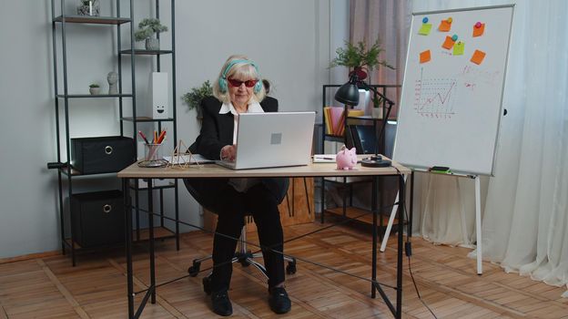 Senior business woman wears sunglasses, listening music on headphones at workplace desk dancing victory dance, weekends. Rejoicing developing project. Celebrating success. Motivated people in office