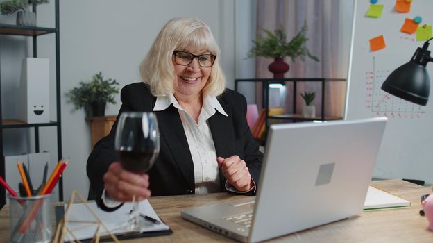 Senior business woman freelancer gets good news, positive exam results, opportunity, successfully signed contract, celebrates victory drinking wine after working on laptop computer sitting at office