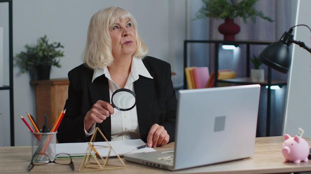 Accountant senior business woman looks through magnifying glass, investigate, examine analyzing reading documents, sits at workplace desk. Elderly grandmother freelancer working at office. Paperwork
