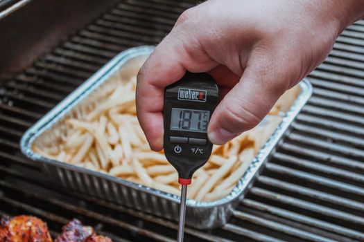 Gatineau, Quebec Canada - July 18, 2020: A food thermometer checking that chicken is being cooked correctly, safely on a barbecue along with Fried potato. Fries on a foil pan. Weber thermometer