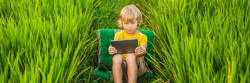 Happy child sitting on the field holding tablet. Boy sitting on the grass on sunny day. Home schooling or playing a tablet. BANNER, LONG FORMAT