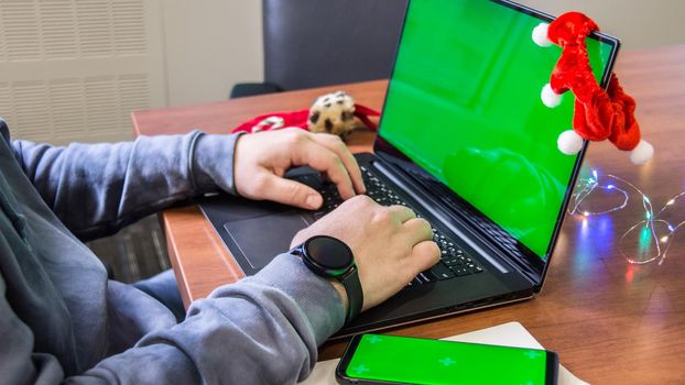 Closeup man hands typing on laptop keyboard with empty green screen, smartphone mockup and Christmas decoration on background. Holiday concept. Office. Mock-up.