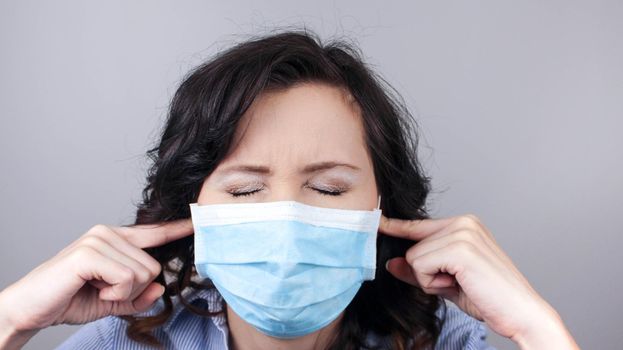 Portrait of nervous brunette caucasian young woman with surgical medical mask standing and putting finger on ears and don't want to hear. studio shot, isolated on grey background.