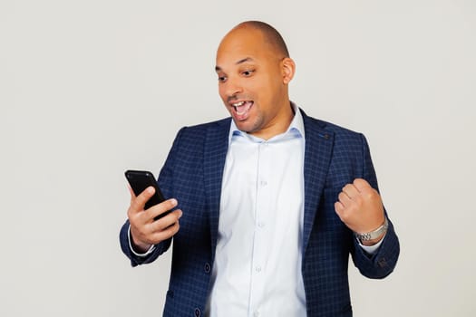 Portrait of happy young african american businessman guy using smart phone, screaming with pride and celebrating victory and success, very excited, rejoicing emotions. Standing on a gray background.
