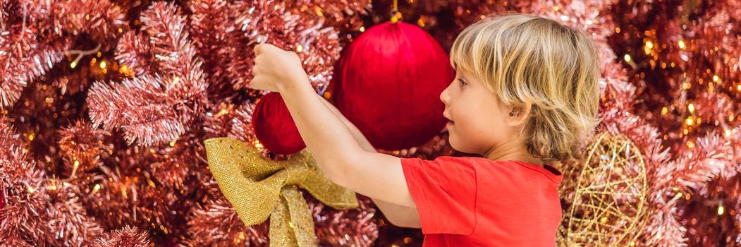 Boy hangs a decoration on the red Christmas tree. Red Christmas tree on the background of lights. BANNER, LONG FORMAT