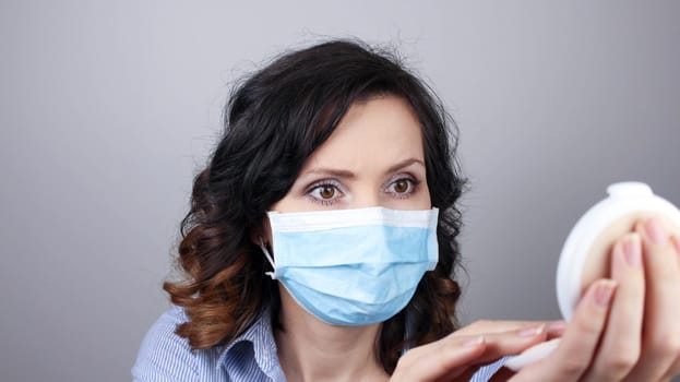 Woman wearing protection face mask against coronavirus and applying make-up. Woman in a mask looking in a mirror. Medical mask, Close up shot, Select focus, Prevention from covid19