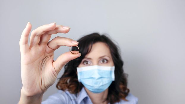 Woman in protective mask holding wedding ring against light background, focus on hand. Divorce during coronavirus quarantine