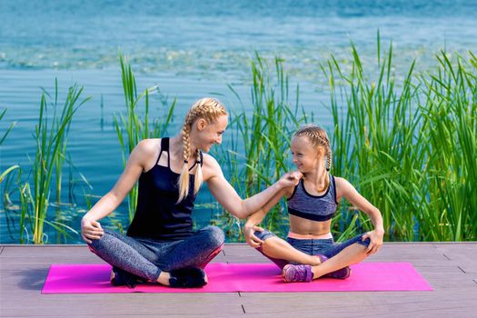 Portrait of mother and daughter sitting on yoga mat by the lake outdoors. Healthy and exercise concept.