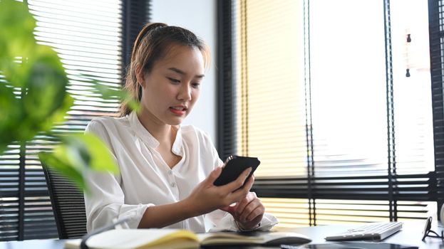 Asian female manager sitting at her workplace and using mobile phone for communicating in social network.