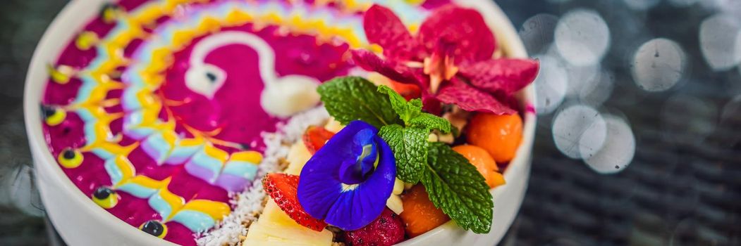 BANNER, LONG FORMAT Healthy tropical breakfast, smoothie bowl with tropical fruits, decorated with a pattern of colorful yogurt with turmeric and spirulina. It is also decorated with fruits, flowers, chia seeds, coconut, granola, pineapple, mint and strawberries.