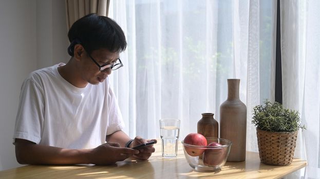 Asian man sitting at kitchen table and using smart phone for checking news or social media.