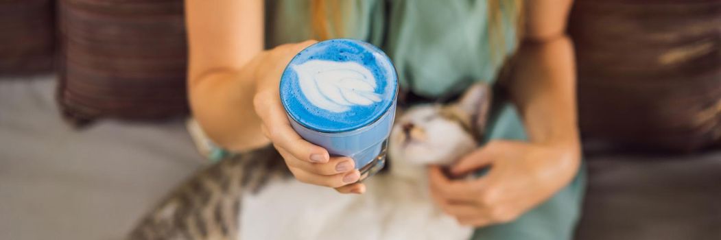 Young woman having a mediterranean breakfast seated at sofa and with her cat and drinks Trendy drink: Blue latte. Hot butterfly pea latte or blue spirulina latte. BANNER, LONG FORMAT