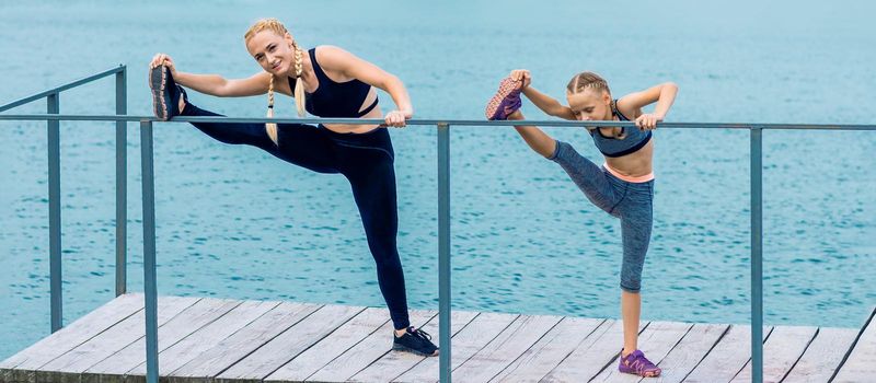 Sportive happy mother is doing workout with little daughter on the shore of the lake.