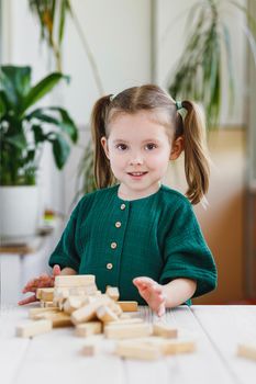 Smiling canny cute child in green dress and broken wooden jenga tower on a table. Indoor activities concept.