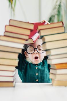 Cute little girl in round shape glasses and green dress behind arch made of books. Reading and kids education concept.
