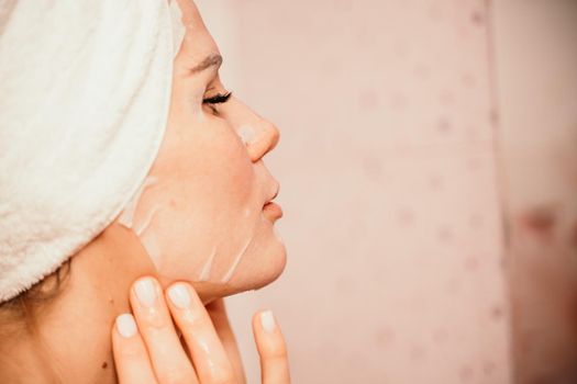 Young beautiful woman using a moisturizing facial mask after taking a bath. Pretty attractive girl in a towel on her head stands in front of a mirror in a home bathroom. Daily hygiene and skin care.