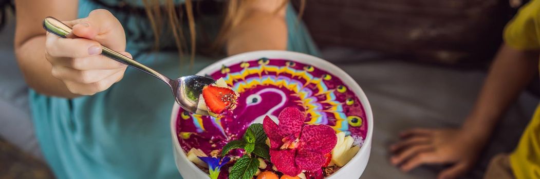 BANNER, LONG FORMAT Mother and son having a mediterranean breakfast seated at the sofa and eats Healthy tropical breakfast, smoothie bowl with tropical fruits, decorated with a pattern of colorful yogurt with turmeric and spirulina. It is also decorated with fruits, flowers, chia seeds, coconut, granola, pineapple, mint and strawberries.