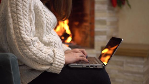 A young pregnant woman is sitting in an armchair by the fireplace and typing on a laptop. The girl writes an email to Santa Claus. 4k