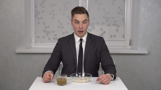 A sleepy young man in a suit eats cereal and drinks coffee and is invigorated. Young businessman at home in the kitchen. 4k