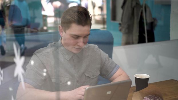 Young man sitting behind the glass in a cafe with a laptop. The man drinks coffee and works. 4k