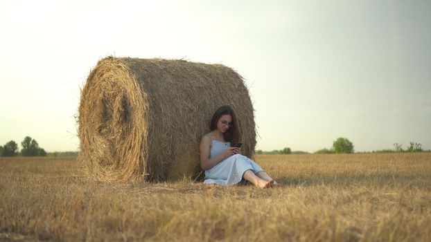 A young woman sits near a haystack in a field with a phone in her hands. A girl with glasses and a blue dress is texting on the phone. 4k