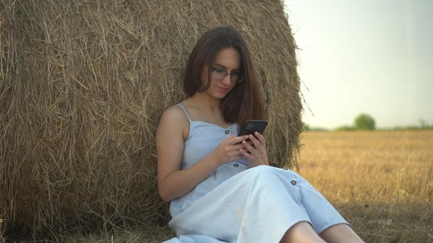 A young woman sits near a haystack in a field with a phone in her hands. A girl with glasses and a blue dress is texting on the phone. 4k
