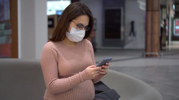 A young pregnant woman in glasses and a medical mask sits with a phone on a bench. Girl in the mall and uses a smartphone. 4k