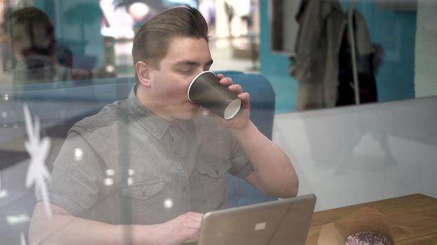 Young man sitting behind the glass in a cafe with a laptop. The man drinks coffee and works. 4k