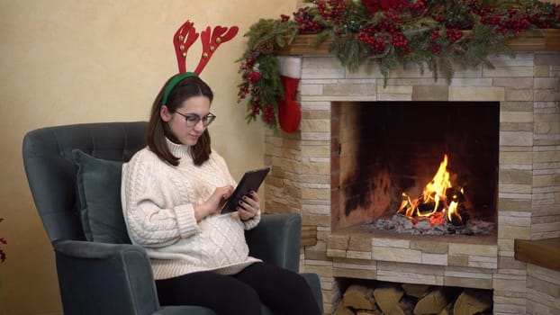 A young pregnant woman with glasses sits in a chair by the fireplace with horns on her head and writes on the tablet. 4k